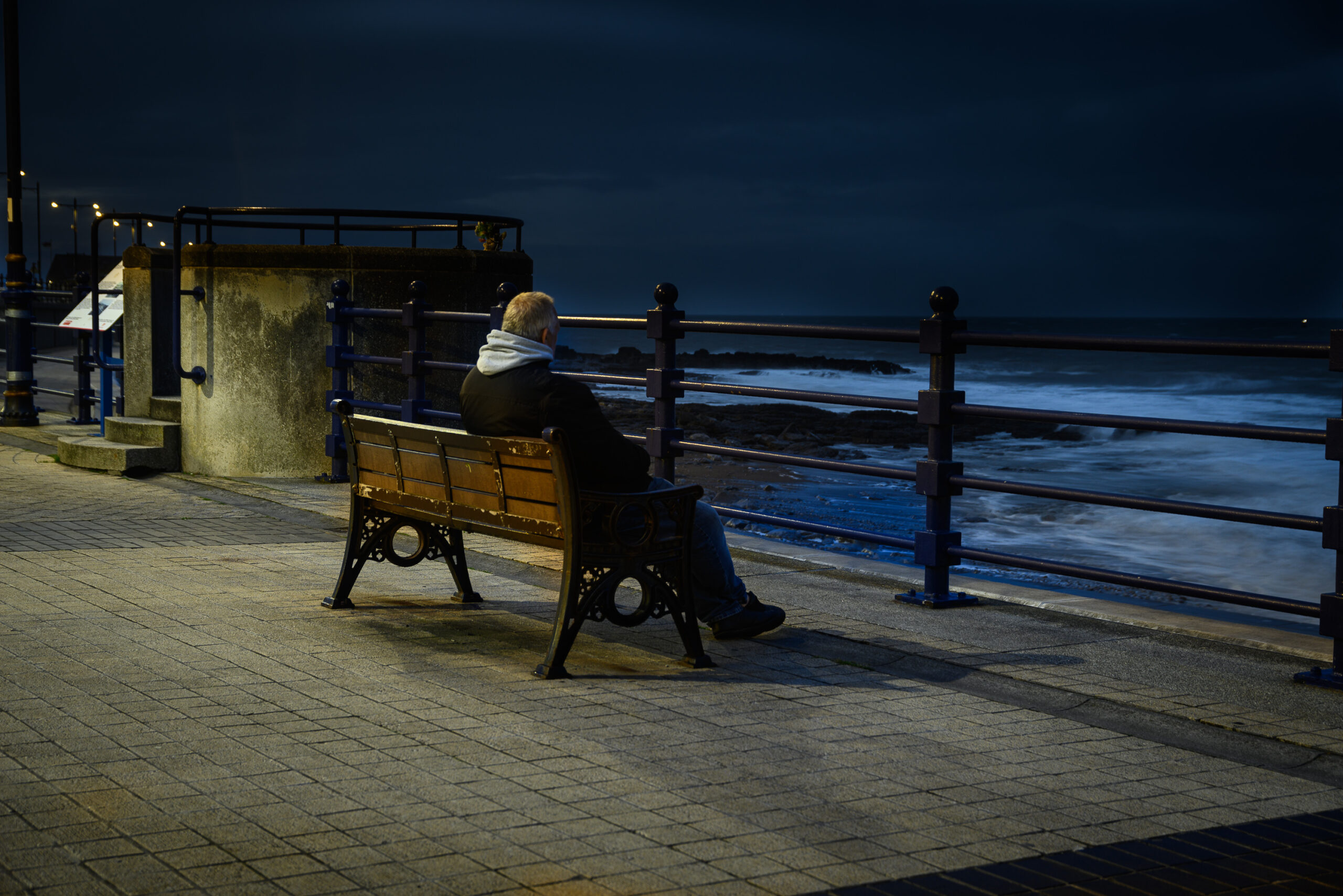 Charlie Hannah cuts a lonely figure, sitting on a bench overlooking a dark, churning sea at night, from the Alone In My Head series by Charlie Hannah ARPS.