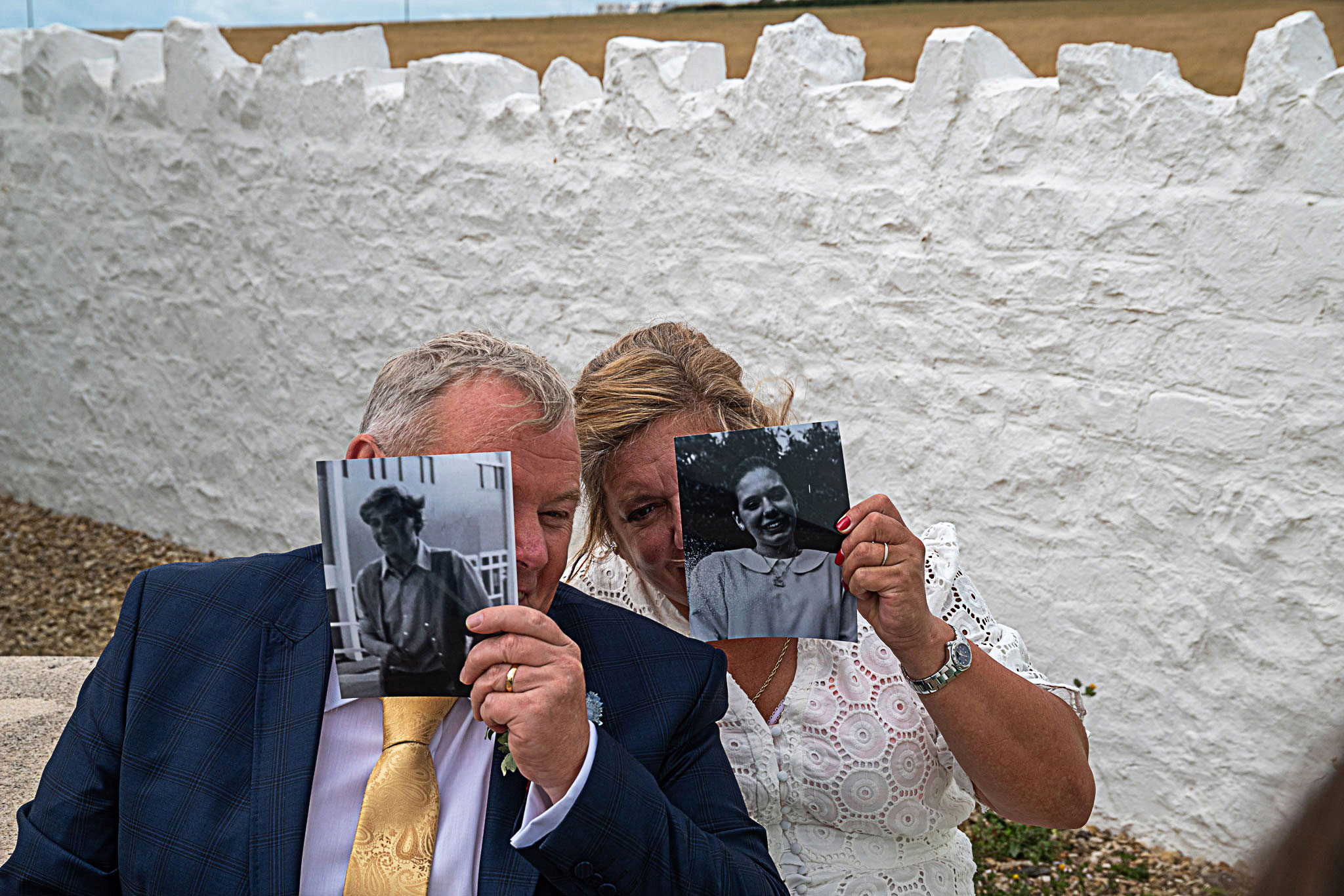 A documentary photograph of a man in a blue suit jacket and gold tie alongside a woman in a white lace dress, both playfully holding up small black and white vintage portraits over parts of their faces in front of a white stone wall.