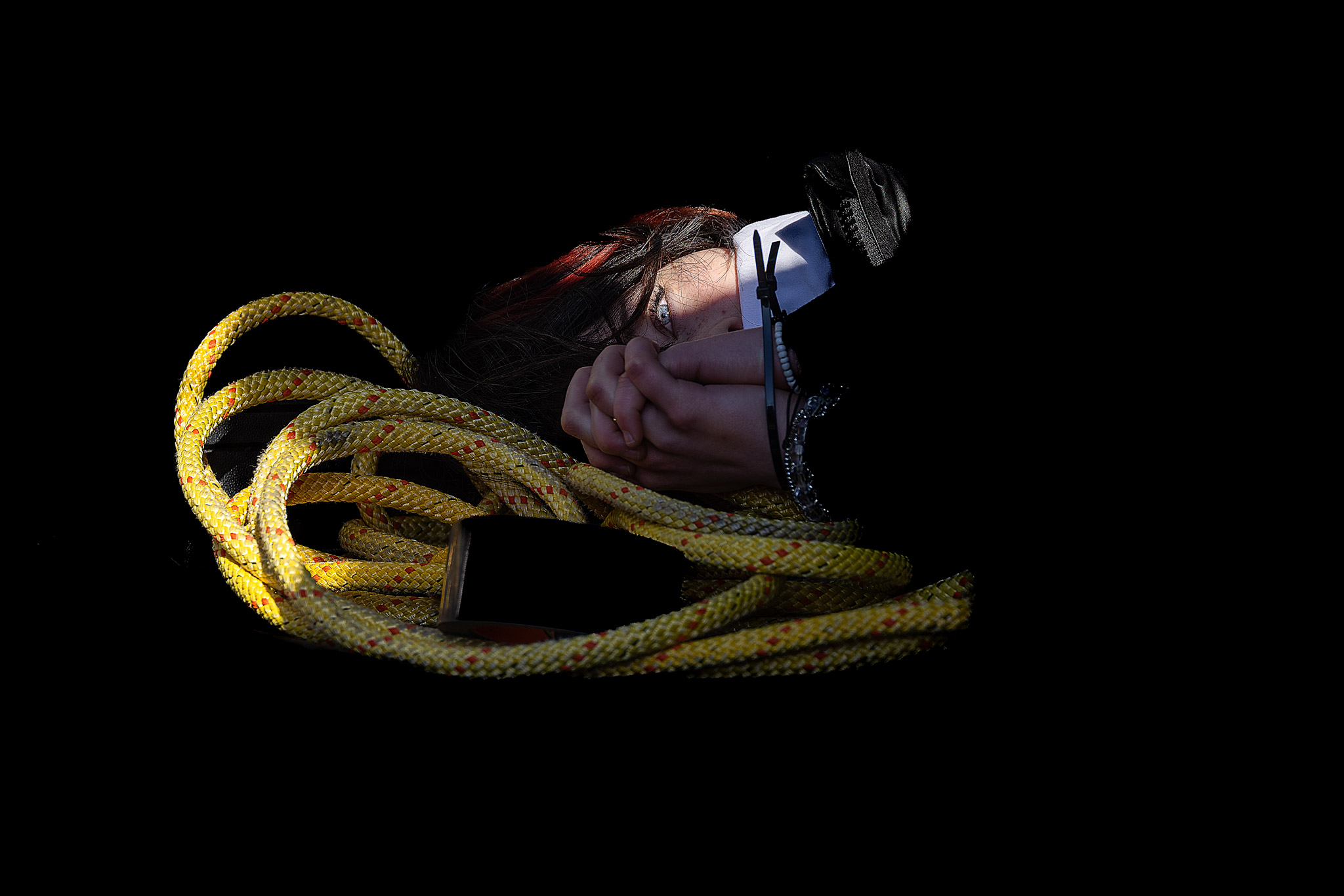 A dramatic fine-art portrait in deep chiaroscuro style, showing a person's hands bound by a black cable tie alongside a coil of bright yellow rope, with one piercing eye peering out from heavy shadows against a completely black background.