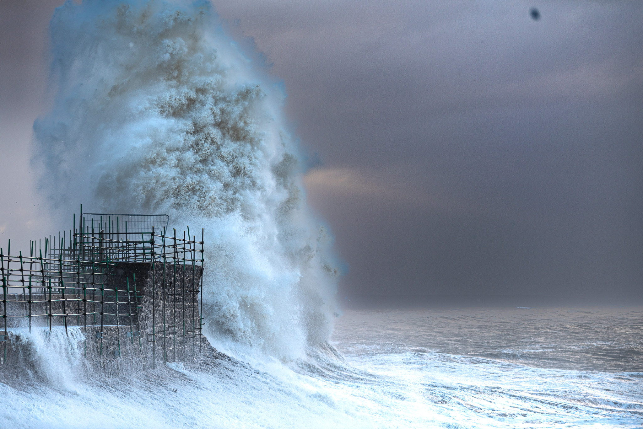 A dramatic seascape photograph of a massive, exploding white ocean wave crashing violently against a stone harbour sea wall covered in scaffolding during a fierce storm under a dark, moody sky in Wales.