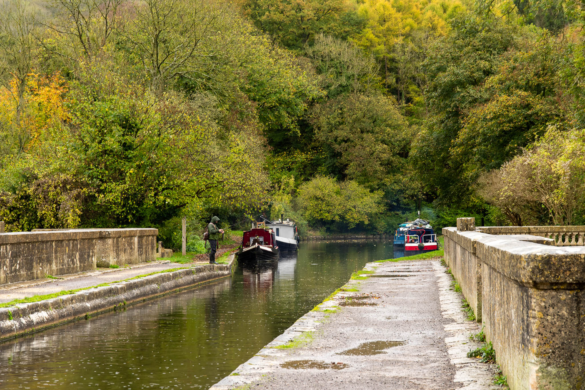 A scenic view looking down a stone canal aqueduct on the kennet and avon canal with narrowboats moored along the water during autumn, featuring a lone angler fishing on the towpath under a dense green and yellow woodland.