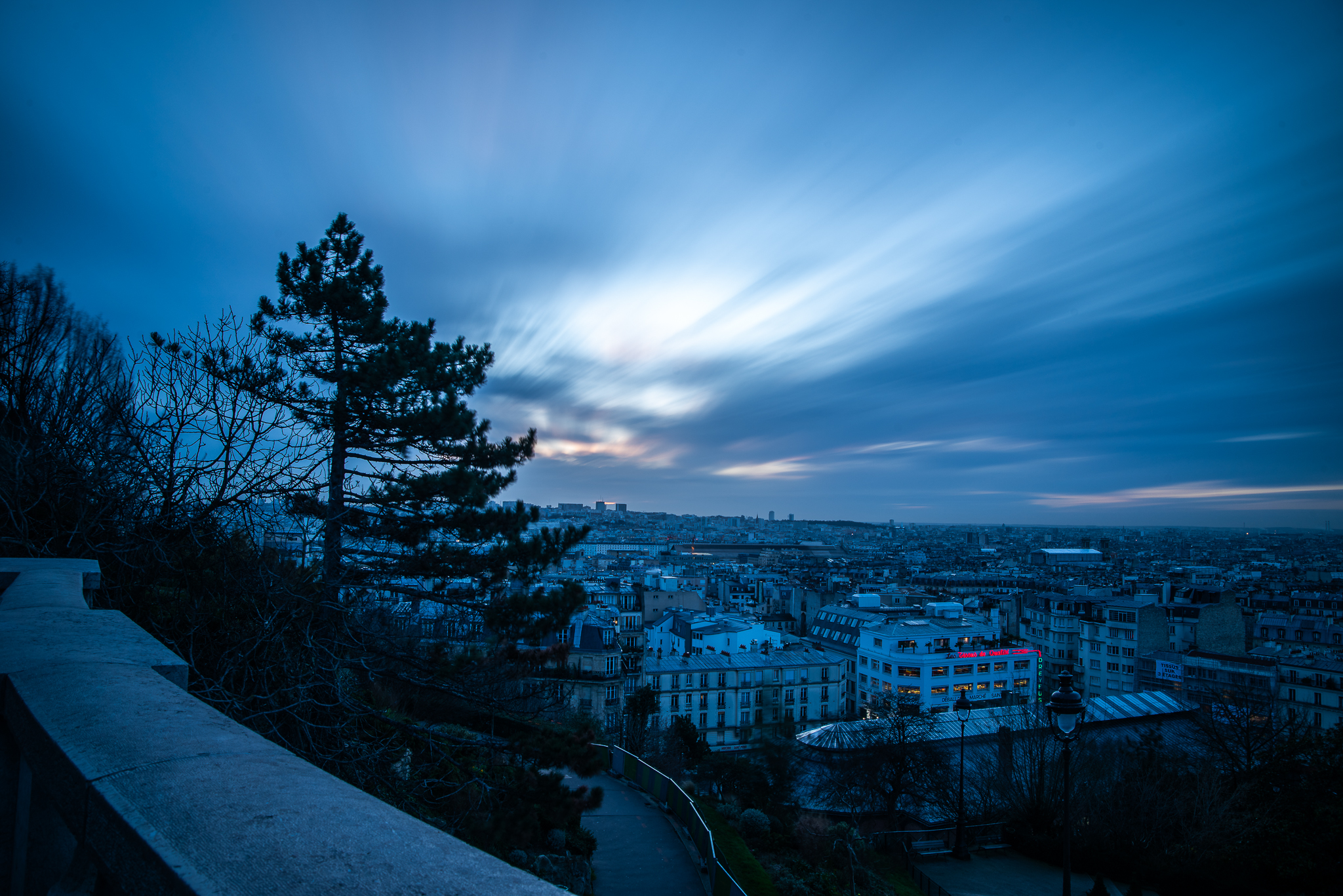 A pre-dawn long-exposure photograph of the sprawling Paris cityscape from Montmartre at blue hour, featuring motion-blurred clouds streaking across the sky, a dark evergreen tree on the left, and distant city lights beginning to glow.