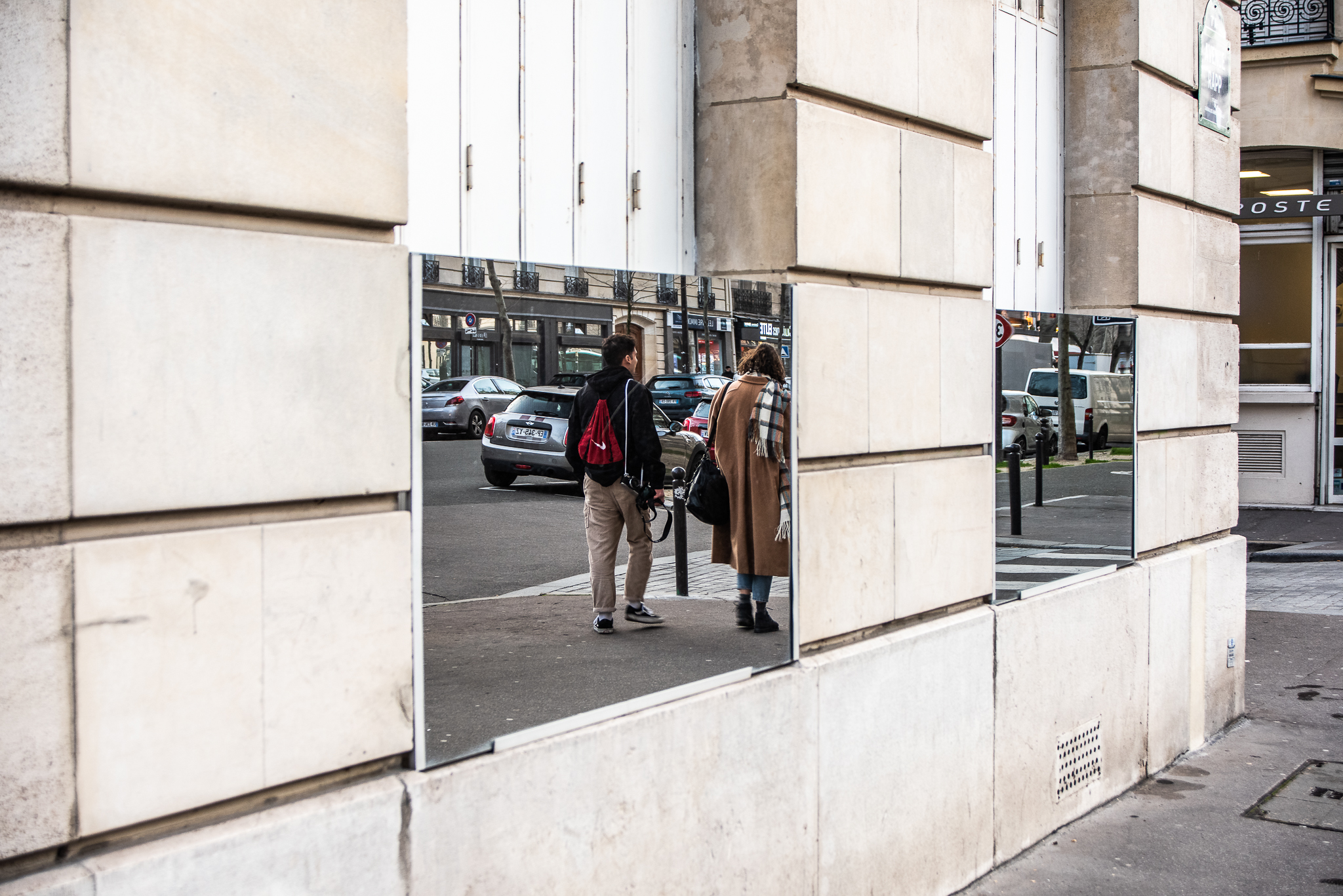 A candid street photograph of two pedestrians walking down a street, captured entirely unawares through rectangular mirrors mounted on a stone building wall in Paris.