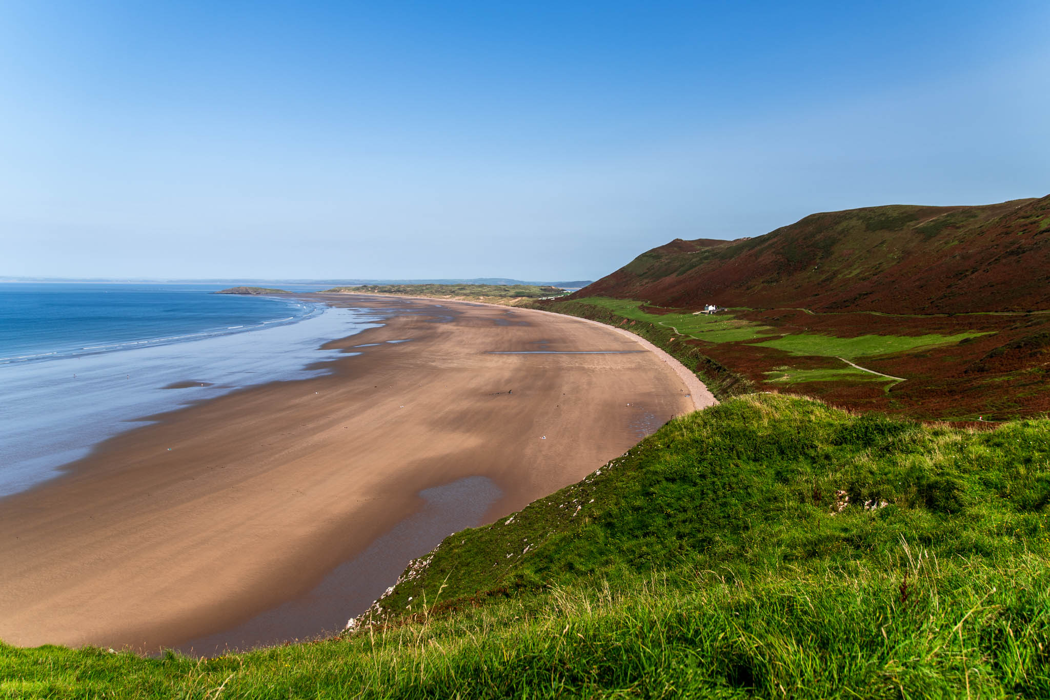 A high-angle landscape photograph looking down at the vast sandy shoreline of Rhossili Bay in South Wales, featuring rolling green and brown cliffs on the right, calm blue ocean waves, and a clear blue sky.