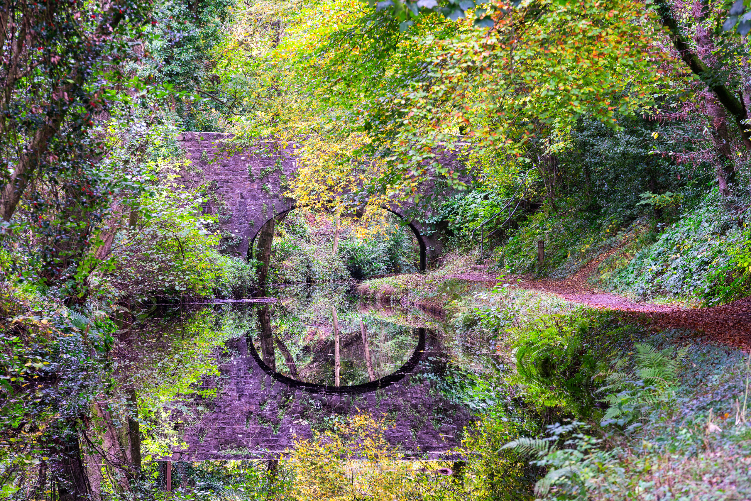 A stone arch bridge over a calm canal in Wales surrounded by lush autumn trees, creating a flawless geometric circle reflection in the still water.