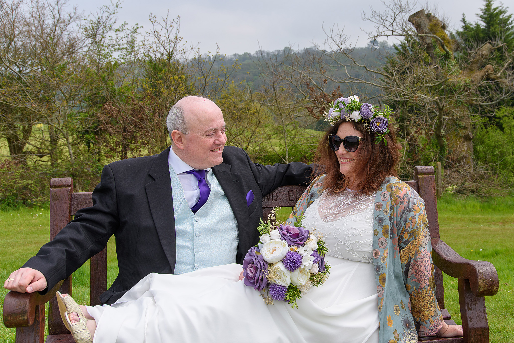 A relaxed bride, Raelene, with her feet up on her groom Gavin's lap, laughing together on a wooden bench outdoors at Tredegar Park Golf Club in Rogerstone, South Wales.
