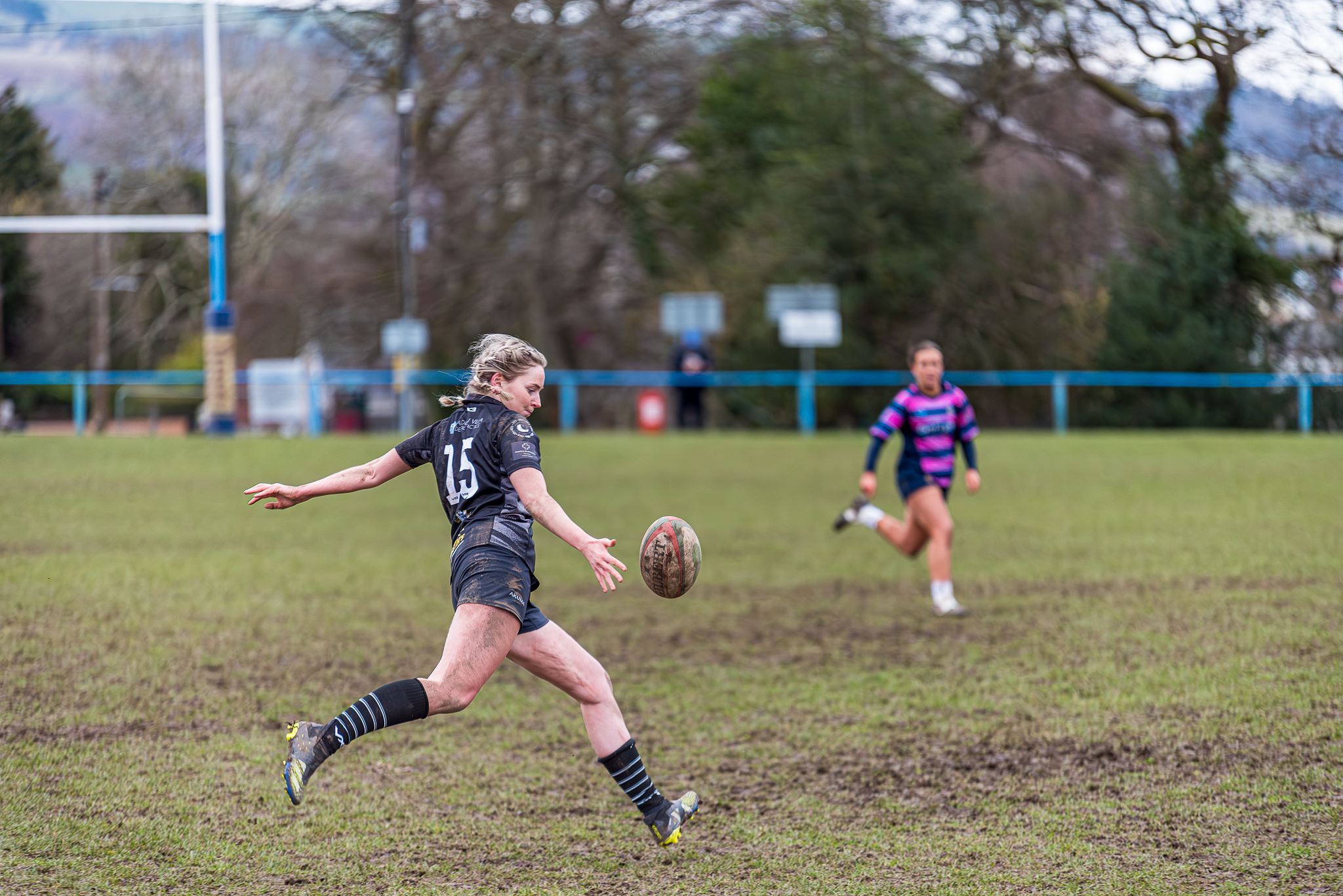 An action sports photograph of a female rugby player wearing a black number 15 jersey kicking a rugby ball on a muddy grass pitch, with an opposing player in a pink and blue jersey running in the background under a shallow depth of field.
