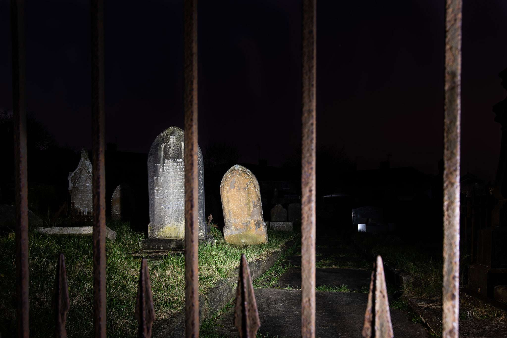 Old gravestones illuminated at night behind rusted iron cemetery gates under a dark sky.