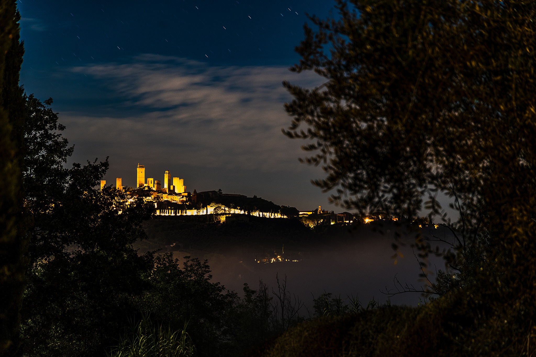 Illuminated medieval towers of San Gimignano glowing against a dark night sky and mist-filled valley framed by trees.