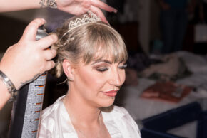 A candid documentary photo of a bride having her tiara adjusted and hair sprayed during final wedding morning preparations.