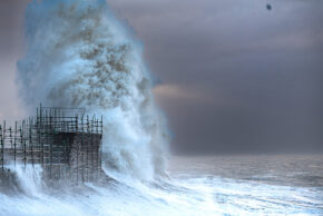 A dramatic view of a massive, exploding white ocean wave crashing violently against a stone harbour sea wall covered in scaffolding during a fierce storm under a dark, moody sky in Wales. Part of the seascape portfolio by Charlie Hannah