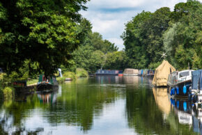 A vibrant summer view looking down a calm canal lined with colourful narrowboats and canal boats moored on both sides, with lush green trees casting deep reflections on the still water under a partly cloudy blue sky.