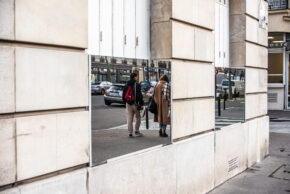 A candid street photograph of two pedestrians walking down a street, captured entirely unawares through rectangular mirrors mounted on a stone building wall in Paris.