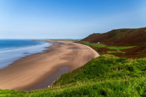 A high-angle landscape photograph looking down at the vast sandy shoreline of Rhossili Bay in South Wales, featuring rolling green and brown cliffs on the right, calm blue ocean waves, and a clear blue sky.