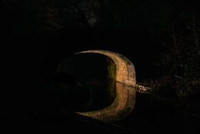 Fine art night photography of an illuminated stone arch bridge reflecting a perfect circle in the dark, still water of the Monmouthshire and Brecon Canal near Llanover.
