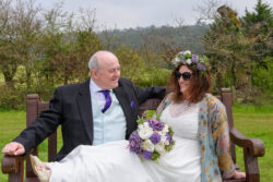 Candid and natural outdoor wedding photography featuring Gavin and Raelene at Tredegar Park Golf Club, captured by South Wales documentary wedding photographer Charlie Hannah. A relaxed bride, Raelene, with her feet up on her groom Gavin's lap, laughing together on a wooden bench outdoors at Tredegar Park Golf Club in Rogerstone, South Wales.