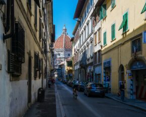 A narrow street in Florence, Italy, framed by tall historic buildings with the dome of the Florence Cathedral visible in the distance under a blue sky.