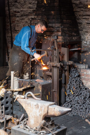 Black Country Living Museum, Dudley - May 2022 Action photography of a traditional blacksmith working hot metal on an anvil with flying sparks inside a historic forge.
