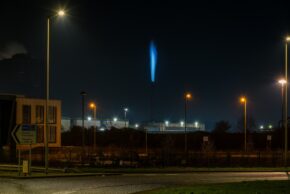 A striking night photograph capturing a brilliant electric-blue industrial flare shooting into the night sky. Framed by sharp streetlamp starbursts and roadside signage, this long-exposure shot highlights the surreal and dramatic glow of heavy industrial activity after dark. A nightscape featuring a vivid electric-blue industrial flare stack burning brightly against a dark sky, framed by streetlamps and roadside infrastructure in the foreground.