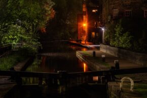 A tranquil canal lock at night with glowing street lamps and historic stone buildings reflecting on the water