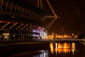 A moody night view of the iconic Cardiff Millennium Stadium reflected in the river below. I know it's the Principality Stadium but back then it wasn't Long exposure night photography of the illuminated Cardiff Millennium Stadium and its structural pylons with warm light reflections shimmering across the river.