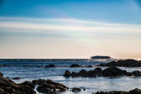 Dark jagged foreground rocks with a long-exposure sea and the blurred motion of a catamaran ferry on the horizon