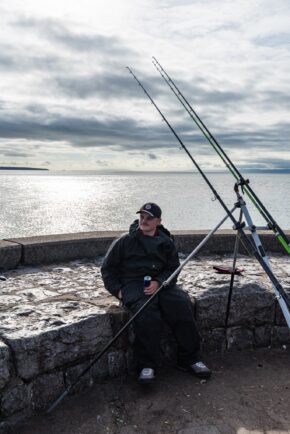 Lifestyle and sports-adjacent outdoor photography capturing an angler on a sea wall, perfectly highlighting the quieter, more patient side of coastal pastimes. An angler sitting relaxed by the sea next to dual fishing rods mounted on a tripod against a coastal backdrop.
