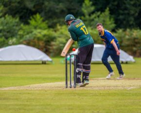 Action sports photography of a cricket batsman wearing number 17 defending the wicket as the ball passes, with a fielder reacting in the background.