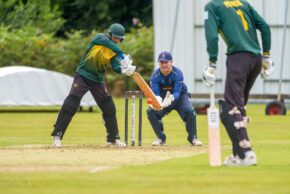 Dynamic sports photography capturing a batsman in green and gold facing the wicketkeeper during a local UK cricket match. Cricket batsman playing a defensive stroke with the wicketkeeper and umpire ready behind the stumps during a match.