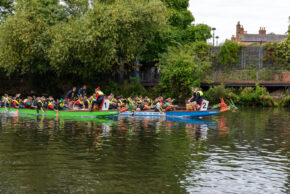 Action-packed sports photography capturing rival dragon boat crews racing side-by-side on a calm river surrounded by trees. Two dragon boat teams competing head-to-head, paddling furiously across the water during a local regatta.