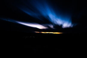 Beddau - February 2020 Long exposure night sky photography showing dramatic streaks of blue clouds stretching across a dark sky above a glowing horizon and silhouetted hills.