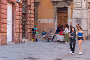 Street photography capturing local life with a street vendor and pedestrians walking across a historic stone square in Perugia, Italy.