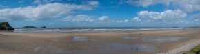 A wide panoramic view of the expansive sandy expanse of Rhossili Beach and Worms Head under a blue sky in the Gower Peninsula