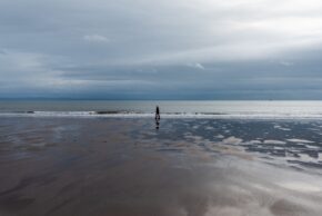 A lone figure walking along a reflective wet sand beach under a vast moody sky