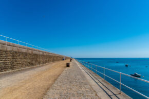 The long granite wall and white railings of St Catherine's Breakwater stretching out into the blue sea in Jersey