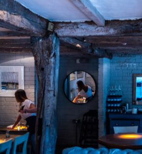A woman reflected in a round mirror inside a rustic pub interior with exposed wooden beams and blue lighting.