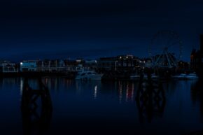 Night view of a calm harbour featuring docked boats, illuminated waterfront buildings, and a Ferris wheel reflecting on the water.