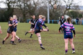 Action sports photography of a women's rugby player kicking the ball downfield during a match on a muddy pitch, watched by teammates and opponents