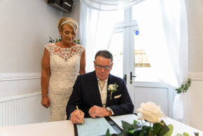 Sharon standing and smiling as Andrew signs the wedding register at Glyn Clydach Hotel in Neath.