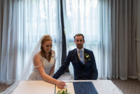 Bride signing the wedding register with the groom sitting beside her smiling at the camera.