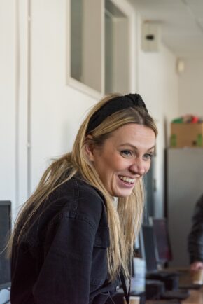 Behind the scenes shot. Candid portrait of a smiling female crew member on set, wearing a black jacket and black headband, looking off-camera.
