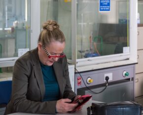 Donna Edwards on set Candid portrait of Welsh actress Donna Edwards seated indoors in a laboratory or workspace setting, focused intently on her phone in a red case.