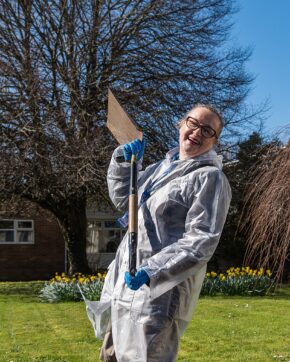 Donna Edwards on the set of Miss Caradog is a Serial Killer Outdoor action portrait of Welsh actress Donna Edwards wearing a white protective coat and blue gloves, holding up a spade with a joyful expression.