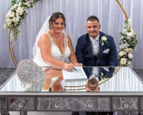 Geraldine signing the wedding register while Stephen looks on and smiles beside her at a mirrored table.