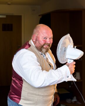 Groom smiling humorously while holding up a small white electric desk fan to cool off indoors on his wedding morning.