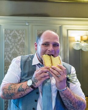 A smiling, tattooed groom in a grey vest and blue tie playfully taking a large bite out of a huge slice of sponge wedding cake at his reception.