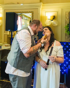 A playful groom feeding a piece of wedding cake to his laughing bride who holds a cake knife at their evening reception party