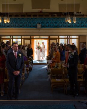 The groom looks on from the front aisle as his bride and her daughter stand framed in the sunlit doorway during the wedding processional.