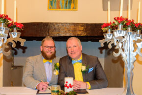 Two grooms smiling at the camera while signing the wedding register together in front of a rustic fireplace.