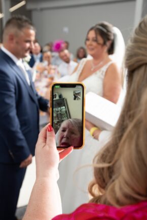 A wedding guest uses a smartphone to video call the bride's grandmother, allowing her to watch the ceremony live while the bride and groom stand nearby.